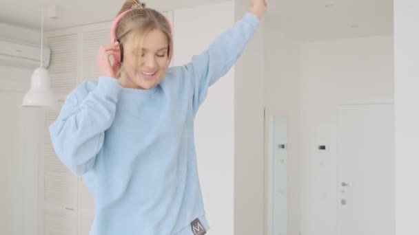 A happy cheerful young blonde woman is enjoying listening the music using headphones in the living room at home - Πλάνα, βίντεο