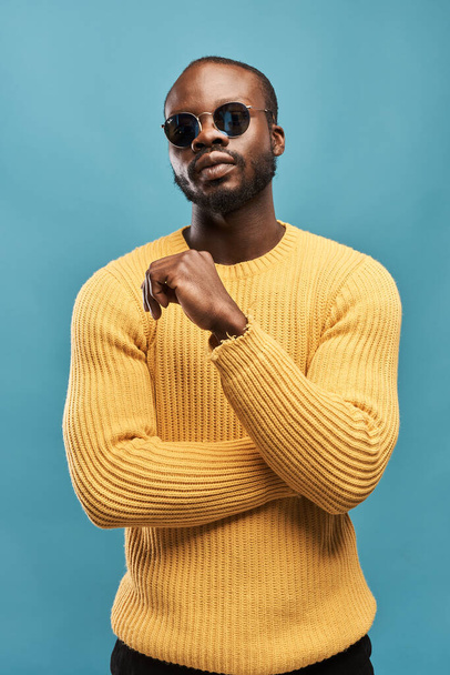 dark-skinned handsome guy with a beard on a blue background in a yellow sweater and sunglasses posing to the camera - Foto, Imagen
