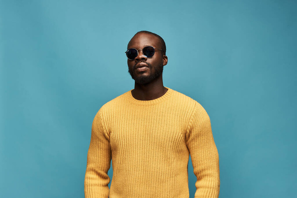 dark-skinned handsome guy with a beard on a blue background in a yellow sweater and sunglasses posing to the camera - Photo, Image