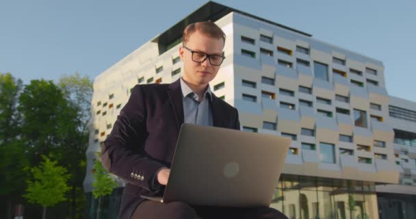 Egy üzletember befejezi a munkát egy laptopon. Egy férfi ül egy parkban egy üzleti központ közelében. Egy mosolygós férfi szemüveggel. 4K - Felvétel, videó