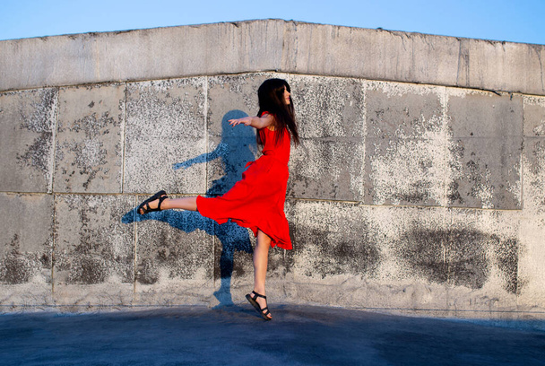 Wonderful long-haired girl dancing outside. Full length view of joyful young woman standing wear in red dress on one leg. - Photo, Image