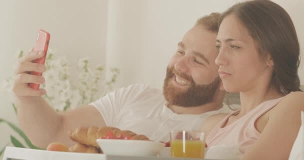 Close up of smiling couple doing selfies and looking at photos while lying on bed early morning. Cheerful man and woman using having fun during breakfast in bedroom. Concept of leisure. - Πλάνα, βίντεο