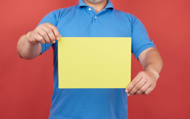 mężczyzna w niebieskich ubraniach trzyma pusty arkusz żółtego papieru na czerwonym tle, miejsce na napis - Zdjęcie, obraz