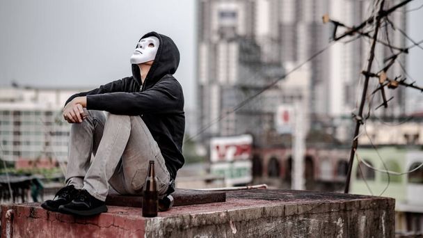 Μυστηριώδης άνθρωπος με λευκή μάσκα με μπουφάν hoodie κάθεται με σπασμένο μπουκάλι αλκοολούχο ποτό σε εγκαταλελειμμένο κτίριο αισθάνεται κουρασμένος και τόνισε. Κατάθλιψη, πρόβλημα ψυχικής υγείας ή αλκοολισμός - Φωτογραφία, εικόνα