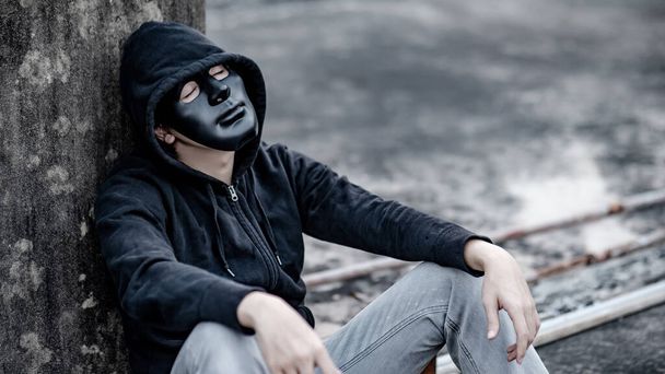 Rätselhafter Mann mit schwarzer Maske und Kapuzenjacke sitzt müde und gestresst auf einem verlassenen Gebäude. Depressionen (schwere depressive Störung) oder Konzepte zur psychischen Gesundheit.  - Foto, Bild