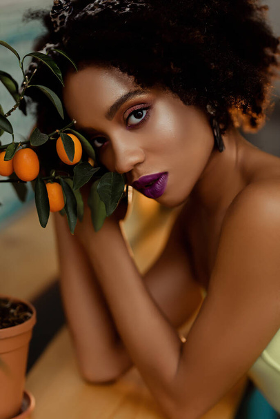 Portret afro-amerykańskiej pięknej młodej kobiety z drzewem pomarańczowym, pięknej kręconej czarnej kobiety z jasnym makijażem, mulatki patrzy w kamerę, miłej pani w gorący letni dzień - Zdjęcie, obraz