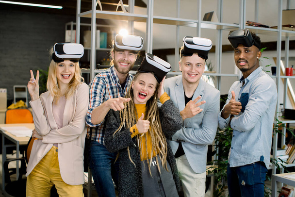 Five cheerful multiethnic friends coworkers enjoying their work with VR goggles at modern creative office room. Young people posing with vr headset smiling and gesturing to camera - Foto, afbeelding