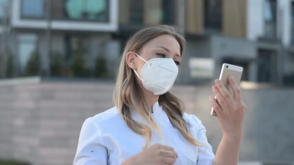 young girl doctor in a protective mask looks at the phone and straightens hair on the street of his city - 映像、動画
