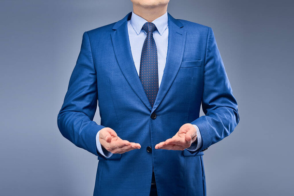 A businessman in a blue suit on a gray-blue background holds his hands in front of him, palms up. Layout for subsequent design. Concept of presentation of goods, services or housing. - Photo, Image