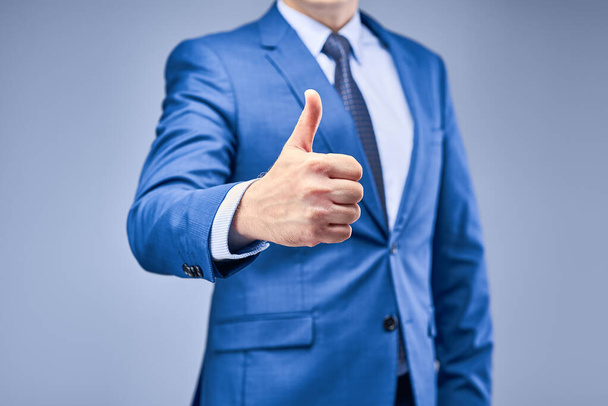A businessman in a blue suit on a gray-blue background holds his hand in front of him in a sign of approval. Hand with thumb up close-up. The concept of approving a decision. - Foto, Imagem