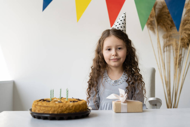Красива дівчина 6-7 років відзначає свій день народження. Вона сидить і перед нею стоїть стіл. Має торт на день народження і невелику коробку з подарунком. Кімната прикрашена для свята. Він легкий і зручний. Концепція свята на
  - Фото, зображення