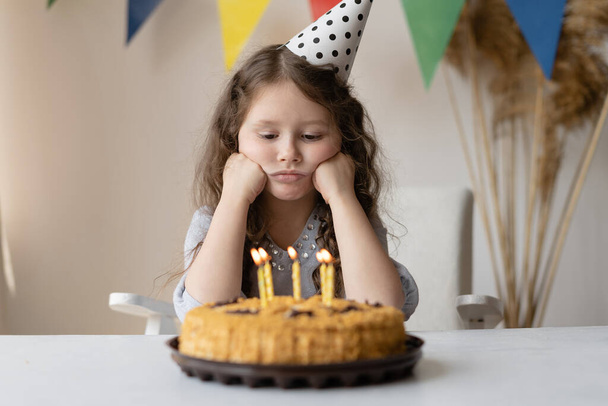 Симпатичный малыш на свой день рождения сидит за столом, подпирая голову двумя кулаками. Перед ней торт со свечами. У нее день рождения и нет настроения. Ребенок расстроен и грустит. Светлая комната и праздничная атмосфера
. - Фото, изображение