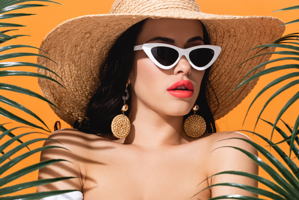 attractive girl in sunglasses and straw hat near palm leaves isolated on orange - Photo, Image