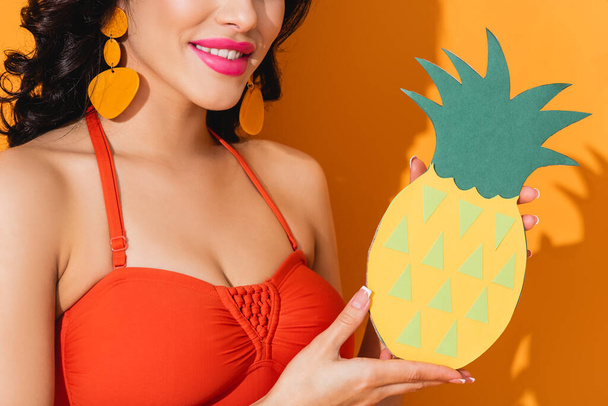 cropped view of happy woman in swimsuit holding paper cut pineapple on orange  - Fotoğraf, Görsel