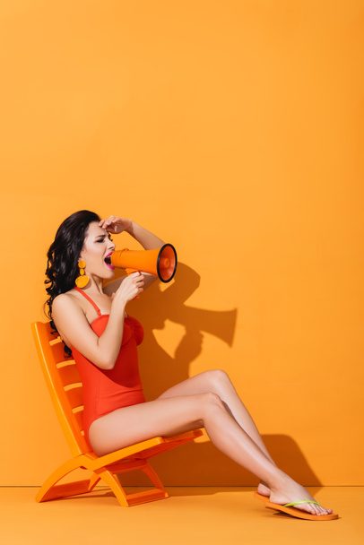 mladá žena v plavkách drží megafon a křičí, zatímco sedí na palubě židle na oranžové - Fotografie, Obrázek