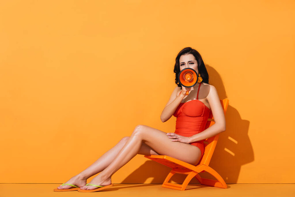 woman in swimwear holding megaphone and screaming while sitting on deck chair on orange - Zdjęcie, obraz