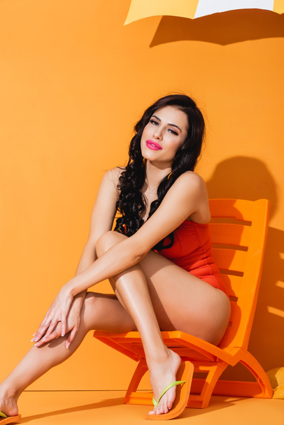 cheerful woman in swimsuit sitting on deck chair on orange - 写真・画像