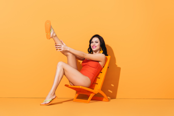 cheerful woman in swimsuit touching leg and sitting on deck chair on orange - Fotoğraf, Görsel