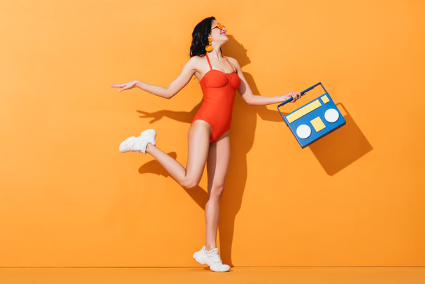 Ευτυχισμένη γυναίκα με αθλητικά παπούτσια, γυαλιά ηλίου και μαγιό που κρατάει χάρτινο κουτί σε πορτοκαλί χρώμα - Φωτογραφία, εικόνα