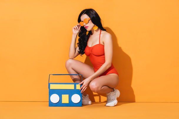 stylish woman in sneakers and bathing suit touching sunglasses while sitting near paper cut boombox on orange - Photo, Image