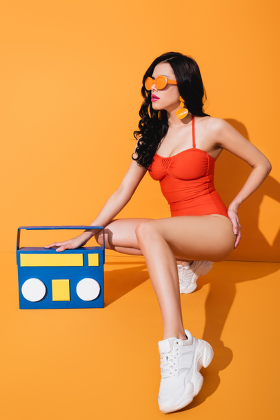 Stylisches Mädchen in Turnschuhen, Badeanzug und Sonnenbrille sitzt neben Papierbastelbox auf orange - Foto, Bild