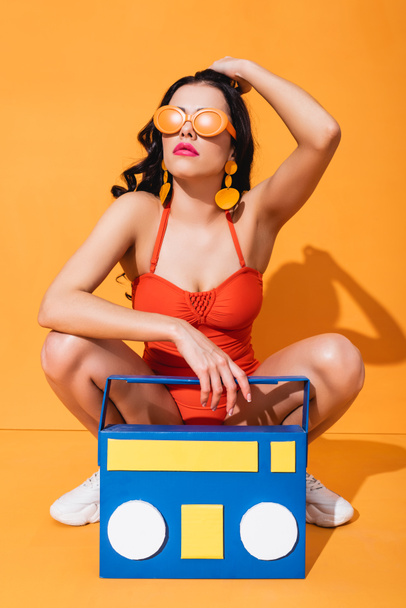 stylish girl in sneakers, bathing suit and sunglasses sitting near paper cut boombox and touching hair on orange - Zdjęcie, obraz