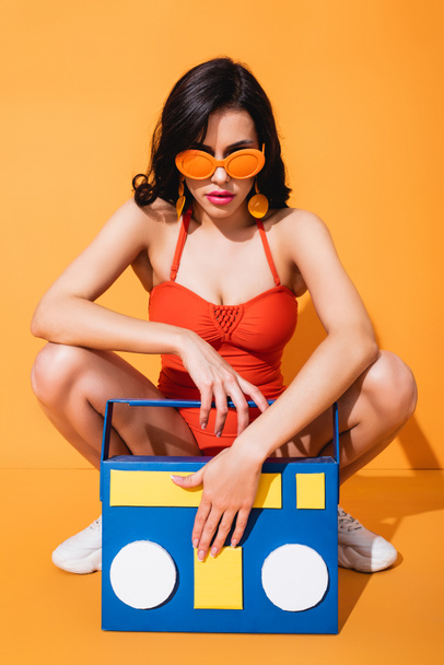 Spor ayakkabılı, mayo giyen ve güneş gözlüklü şık bir kadın. Kağıt kesiğinin yanında oturuyor. Portakallı teyp. - Fotoğraf, Görsel
