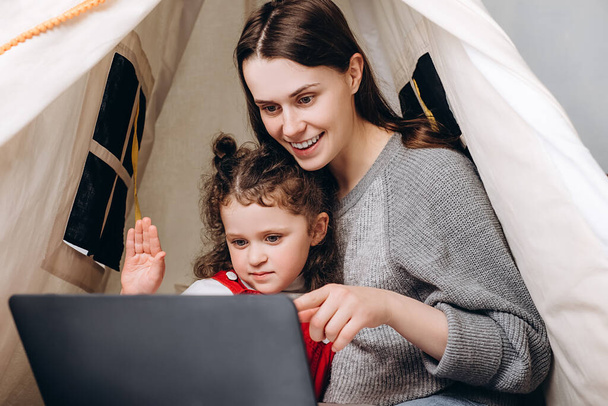 Cheerful young mom with kid daughter waving hands making distance video call looking at laptop sitting in tent at home, loving brunette mother and cute child girl talking to webcamera chatting online - Foto, Imagem