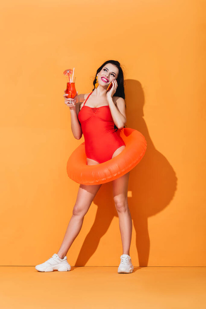 happy girl in swimsuit with inflatable ring holding cocktail and talking on smartphone on orange - Foto, Imagem