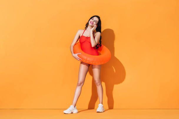 femme heureuse en maillot de bain avec anneau gonflable tenant cocktail et parlant sur smartphone sur orange - Photo, image