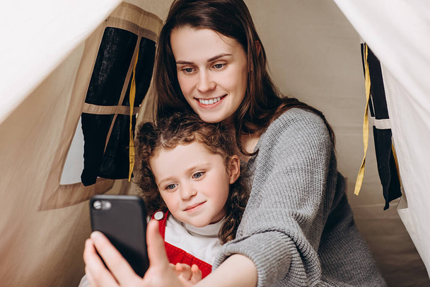 Close up of mum and small girl child relax in tent. Joyful mommy and little daughter sitting together on bed bedding smiling looking at camera feels joyful, making happy selfie photos or call video - Фото, зображення