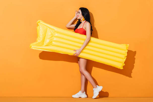 side view of cheerful girl in swimsuit and sneakers holding inflatable mattress on orange  - Foto, Bild