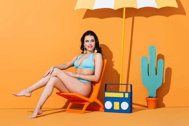happy young woman in swimsuit sitting on deck chair near paper boombox, cactus and umbrella while looking away on orange - Photo, Image
