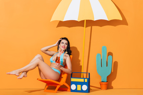 mosolygó nő fürdőruhában ül a fedélzeten szék közelében papír boombox, kaktusz és esernyő, miközben a kezében koktél a narancs - Fotó, kép