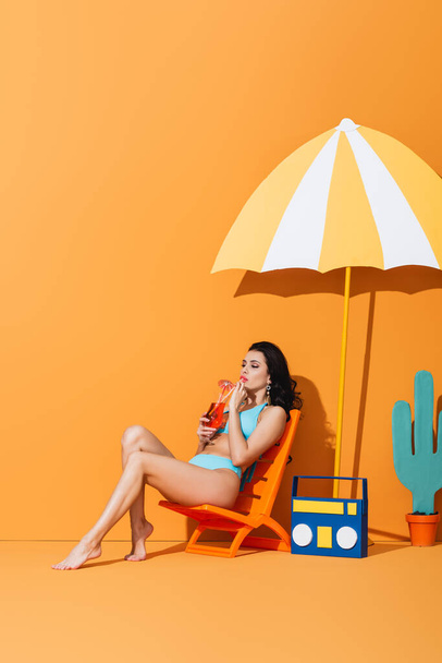woman in swimwear sitting on deck chair near paper boombox, cactus and umbrella while drinking cocktail on orange - Photo, image