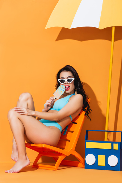trendi nő napszemüvegben és fürdőruhában ül a fedélzeten szék közelében papír boombox és esernyő, miközben nyalogatja fagylalt narancs - Fotó, kép