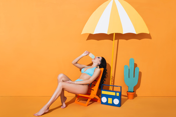 stylish woman in sunglasses and swimwear sitting on deck chair near boombox and umbrella while licking paper ice cream on orange - Foto, imagen