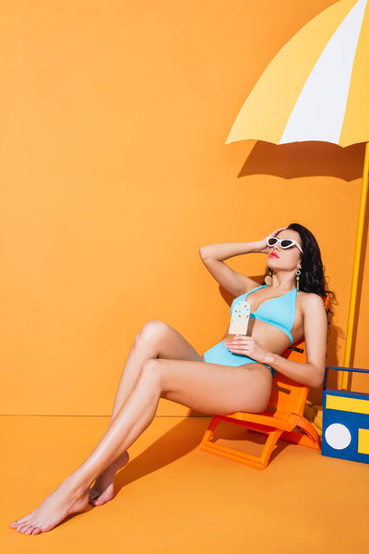 trendy woman in sunglasses and swimwear sitting on deck chair near paper boombox and umbrella while holding ice cream on orange - Foto, imagen