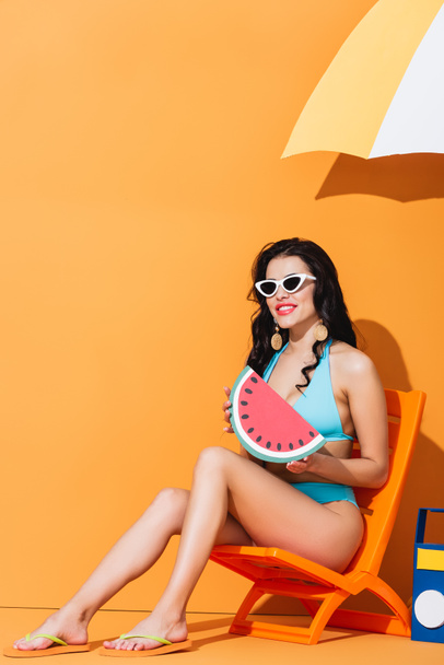 cheerful woman in sunglasses and swimsuit sitting on deck chair near boombox and umbrella while holding paper watermelon on orange - Fotó, kép