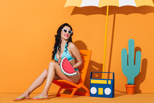 cheerful girl in sunglasses and swimsuit sitting on deck chair near boombox, cactus and umbrella while holding paper watermelon on orange - 写真・画像
