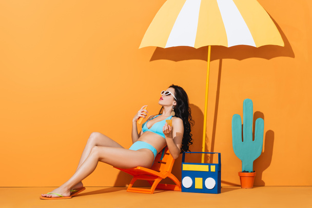 stylish girl in sunglasses and swimsuit sitting on deck chair near paper boombox, cactus and umbrella while applying sunscreen on orange - Photo, Image