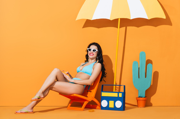 happy girl in sunglasses and swimsuit sitting on deck chair near paper boombox, cactus and umbrella while applying sunscreen on orange - Foto, Imagem