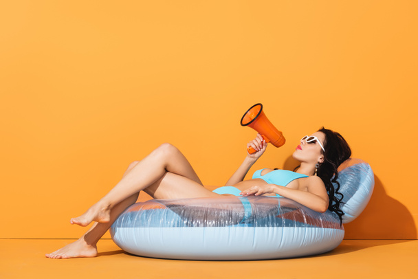 barefoot woman in sunglasses and swimwear lying on inflatable ring and holding megaphone on orange  - Photo, Image