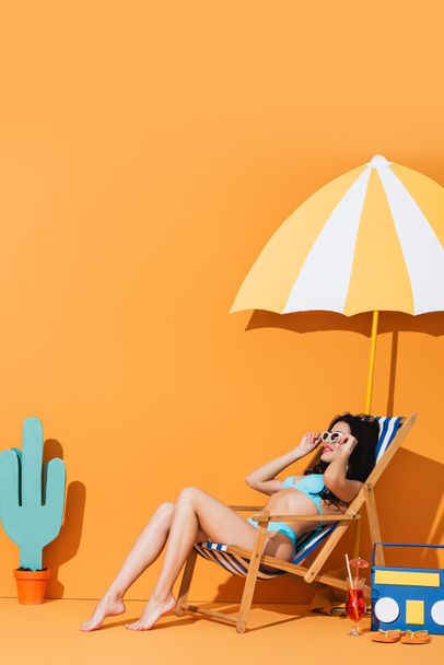 trendy woman in sunglasses and swimwear sitting on deck chair near cocktail, flip flops, paper boombox, cactus and umbrella on orange - Foto, afbeelding