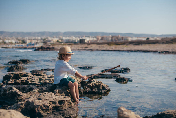 A little boy in a stylish hat sits on a stone and plays with a wand and water. The kid in a fashionable image against the sea sits by the water. Child plays fisherman - Photo, Image