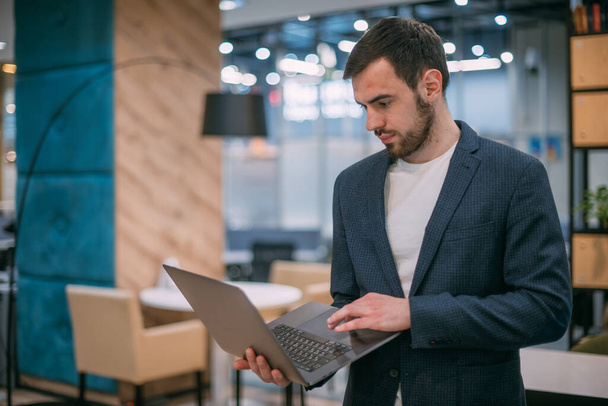 Portret współczesnego młodego biznesmena przy działającym laptopie. Młody przystojny mężczyzna w kurtce we wnętrzu nowoczesnego biura. - Zdjęcie, obraz