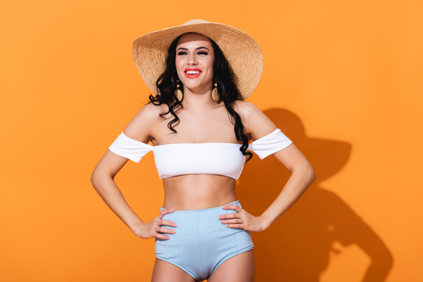 trendy girl in swimsuit standing and straw hat standing with hands on hips on orange  - Foto, afbeelding