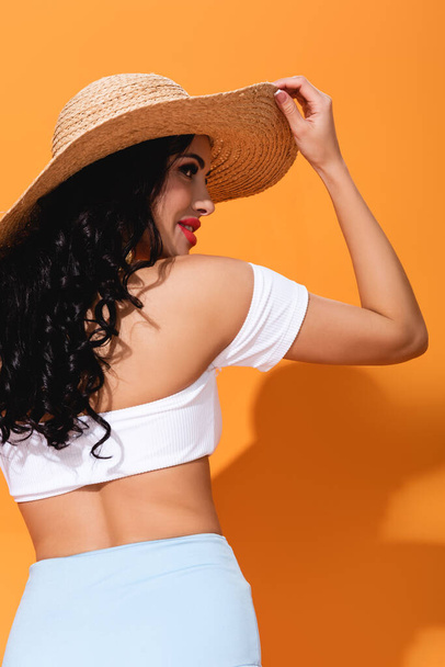 cheerful and stylish girl in straw hat standing on orange  - 写真・画像