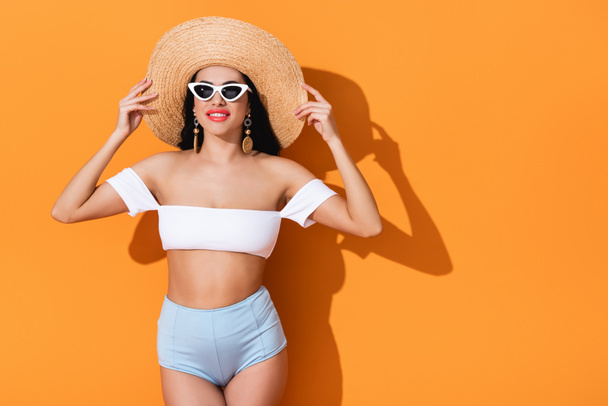 stylish girl in sunglasses smiling and touching straw hat on orange  - Foto, Imagen