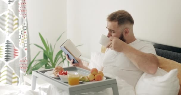 Side view of concentrated guy in middle 30s reading while having breakfast . Handsome bearded man drinking coffee and turning book page while sitting on bed. Concept of leisure. - Filmati, video
